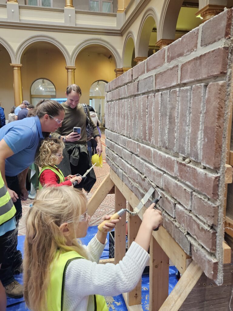 Two children use trowels and pointers to apply mortar to brick wall. NPS employee and smiling parent watch.