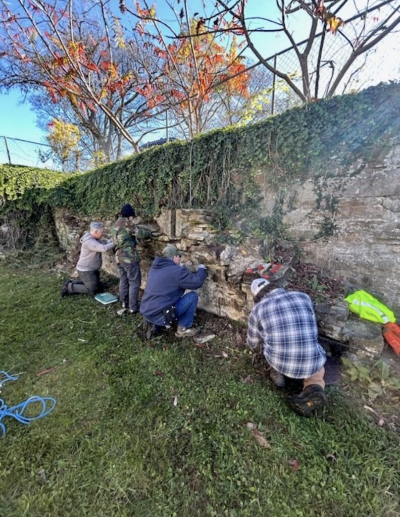 Four attendees of the Arthurdale Heritage masonry workshop repoint a stone retaining wall. Greenery drapes over the top of the wall.